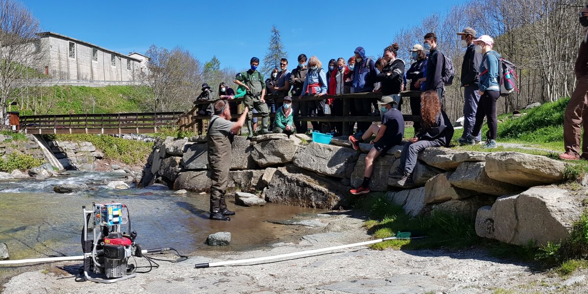 Professor Fenoglio gives a lecture to a group of students during an educational excursion on the Po River in Crissolo. He is seen standing in the water, showing a trout freshly caught using electroanesthesia to the students, who are gathered in a semicircle on the riverbank.