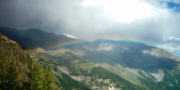 in primo piano un tetto di una abitazione alpina, sullo sfondo un paesaggio montano, con un arcobaleno