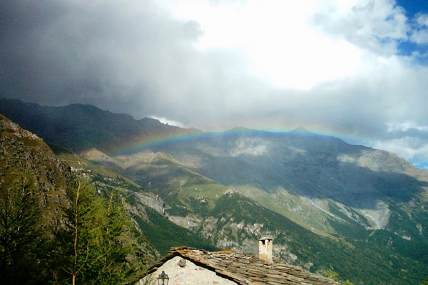 in primo piano un tetto di una abitazione alpina, sullo sfondo un paesaggio montano, con un arcobaleno