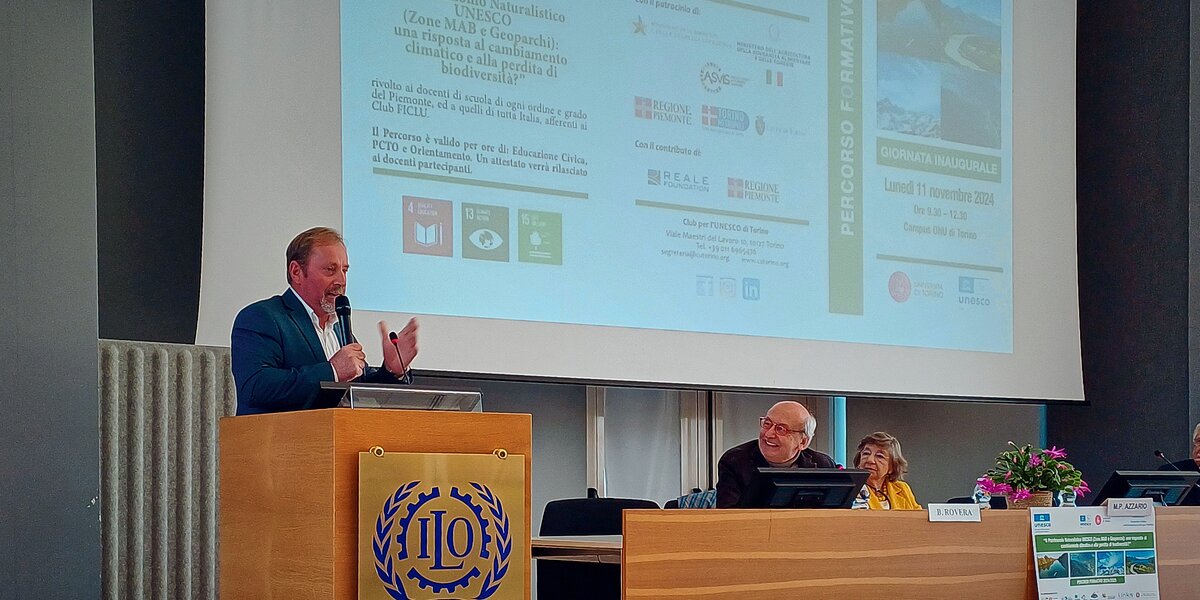 Un momento della presentazione del percorso formativo: il presidente del Parco del Monviso sta parlando, in piedi dietro ad un leggio in legno sulla sinistra della foto, a destra il tavolo dei relatori; sullo sfondo la locandina del corso.