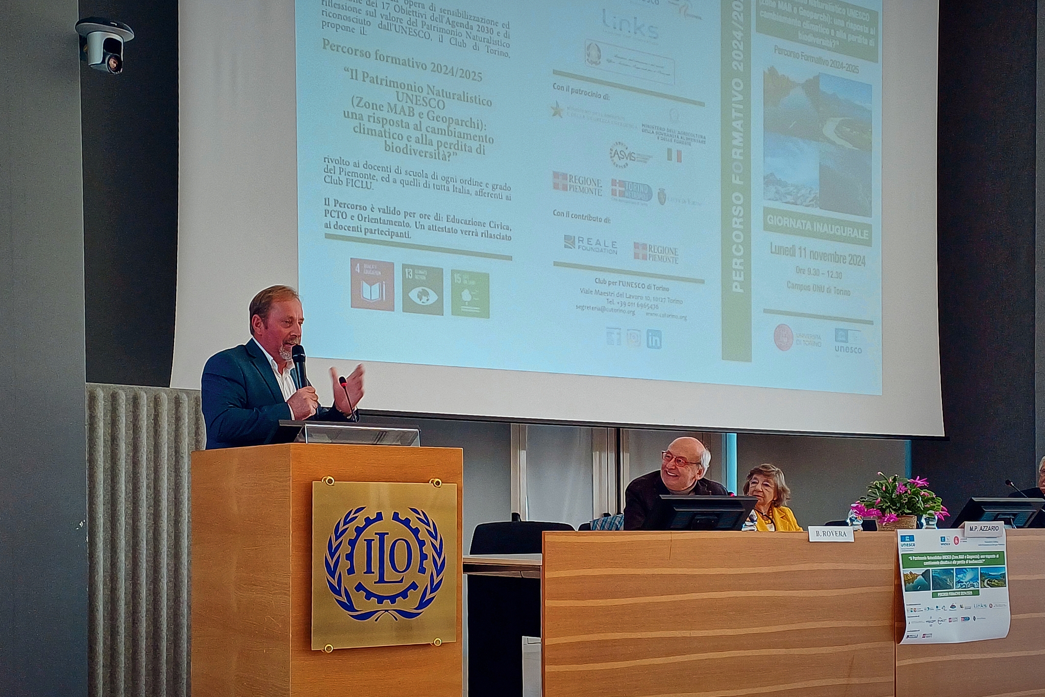 Un momento della presentazione del percorso formativo: il presidente del Parco del Monviso sta parlando, in piedi dietro ad un leggio in legno sulla sinistra della foto, a destra il tavolo dei relatori; sullo sfondo la locandina del corso.