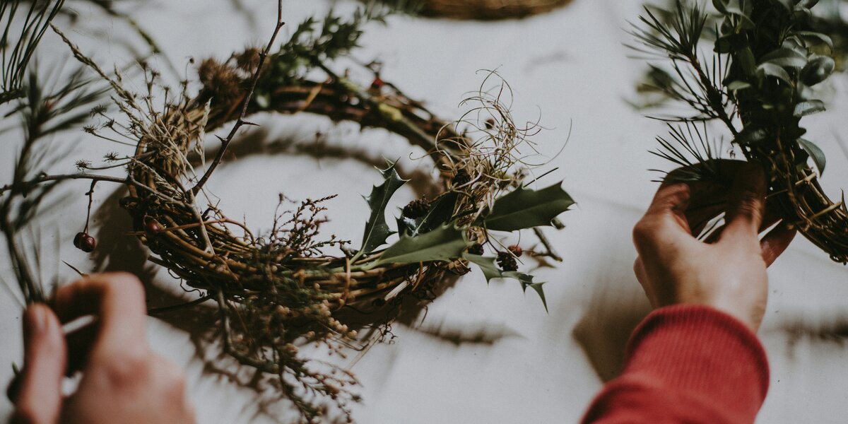 foto ravvicinata di due mani che stanno lavorando su elementi naturali per creare una ghirlanda di Natale: al centro della foto c'è una ghirlanda già pronta