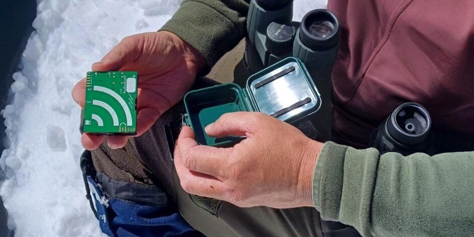 un tecnico del Parco del Monviso mostra un rilevatore bioacustico, una scatoletta verde contenente un chip. Il tecnico è inquadrato dall'alto verso il basso e se ne vedono solo le mani; a fianco ha un binocolo e un monocolo. a terra si vede la neve.