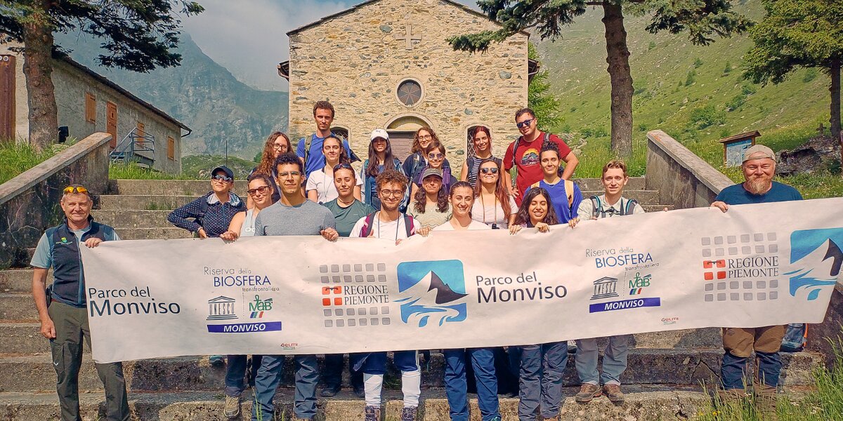foto di gruppo in contesto montano: il gruppo di persone tiene uno striscione con il logo del Parco del Monviso davanti a una chiesetta montana