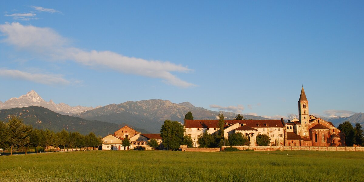 un prato verde e sullo sfondo un complesso di vecchi edifici dove spicca il campanile e il retro della chiesa. dietro le montagne e il monviso
