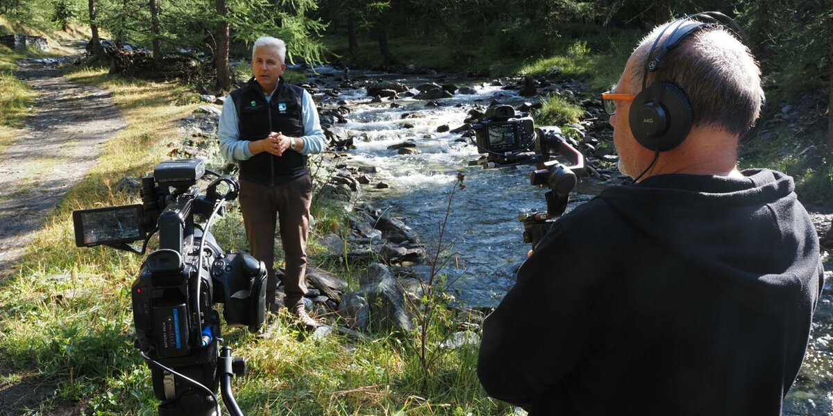 in primo piano un cameraman di schiena sulla destra della foto, sulla sinistra una seconda telecamera su treppiede; sullo sfondo un uomo di circa 60 anni che parla, inquadrato. Il contesto è naturale, a fianco delle persone scorre un torrente e ci sono alberi tutto intorno.