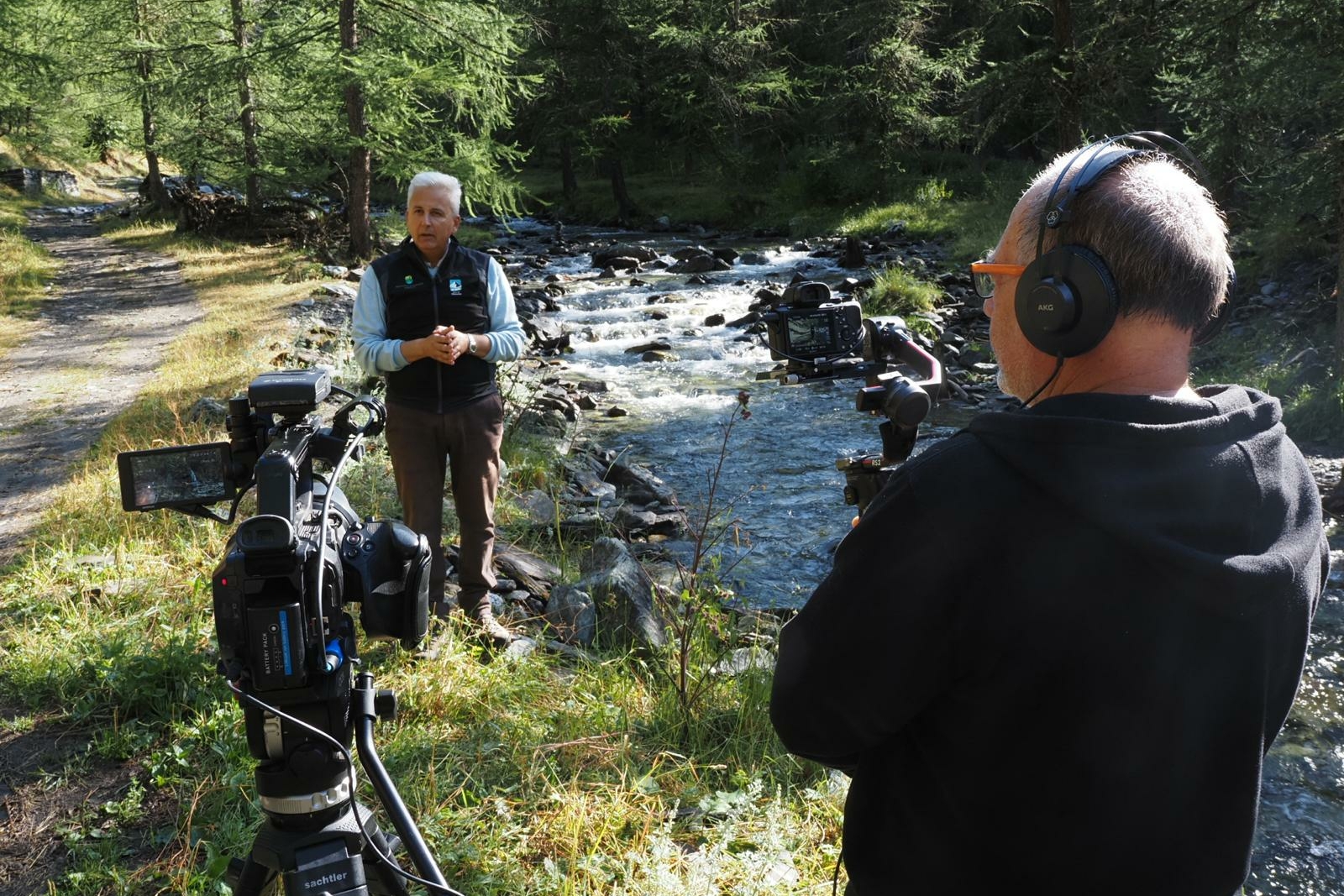 in primo piano un cameraman di schiena sulla destra della foto, sulla sinistra una seconda telecamera su treppiede; sullo sfondo un uomo di circa 60 anni che parla, inquadrato. Il contesto è naturale, a fianco delle persone scorre un torrente e ci sono alberi tutto intorno.