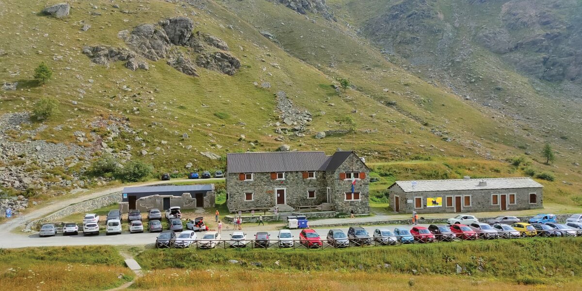 Vista dall’alto di costruzioni situate in una valle montana verdeggiante, la valle Po. Il complesso è composto da due edifici principali in pietra a vista con tetti in lamiera grigia e infissi con persiane marroni: un edificio a due piani più grande e uno più basso a un piano singolo. Davanti agli edifici si estende un ampio parcheggio sterrato dove sono parcheggiate numerose automobili di vari colori disposte in file ordinate. Alcune persone in abiti colorati sono visibili nell'area antistante le costruzioni. Sul lato sinistro è presente una piccola struttura coperta. Alle spalle si ergono ripidi pendii montani erbosi con affioramenti rocciosi grigi e creste frastagliate che raggiungono le quote più elevate. Il paesaggio è tipico delle Alpi occidentali durante la stagione estiva, con vegetazione rigogliosa.