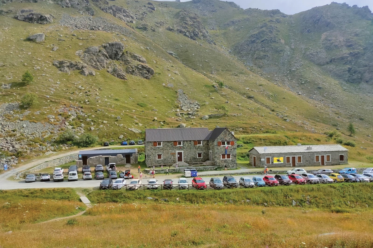 Vista dall’alto di costruzioni situate in una valle montana verdeggiante, la valle Po. Il complesso è composto da due edifici principali in pietra a vista con tetti in lamiera grigia e infissi con persiane marroni: un edificio a due piani più grande e uno più basso a un piano singolo. Davanti agli edifici si estende un ampio parcheggio sterrato dove sono parcheggiate numerose automobili di vari colori disposte in file ordinate. Alcune persone in abiti colorati sono visibili nell'area antistante le costruzioni. Sul lato sinistro è presente una piccola struttura coperta. Alle spalle si ergono ripidi pendii montani erbosi con affioramenti rocciosi grigi e creste frastagliate che raggiungono le quote più elevate. Il paesaggio è tipico delle Alpi occidentali durante la stagione estiva, con vegetazione rigogliosa.
