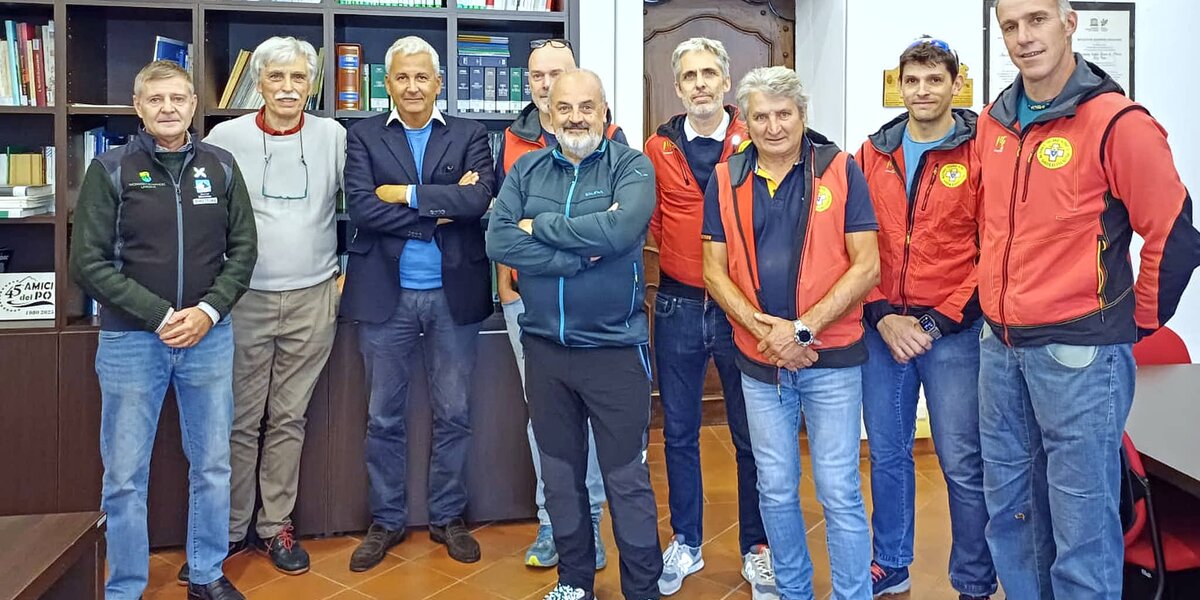 Foto di gruppo di nove uomini in piedi in un ufficio della sede di Saluzzo del Parco del Monviso. Il gruppo è composto da persone di età variabile, prevalentemente di mezza età. Quattro di loro indossano giacche rosse e blu del Soccorso Alpino, riconoscibili dal logo circolare giallo sul petto. Gli altri indossano abbigliamento casual o sportivo: giacche blu, felpe e jeans. Tutti sorridono guardando verso la fotocamera in un'atmosfera cordiale e informale. Sullo sfondo si vede una libreria a scaffali scuri piena di volumi rilegati di vari colori, tra cui spiccano alcuni dorsi verdi allineati. Sulla destra è visibile una porta bianca con un vecchio mobile scuro all'interno e alcune targhe e certificati appesi alla parete. Il pavimento è in piastrelle color terracotta.
