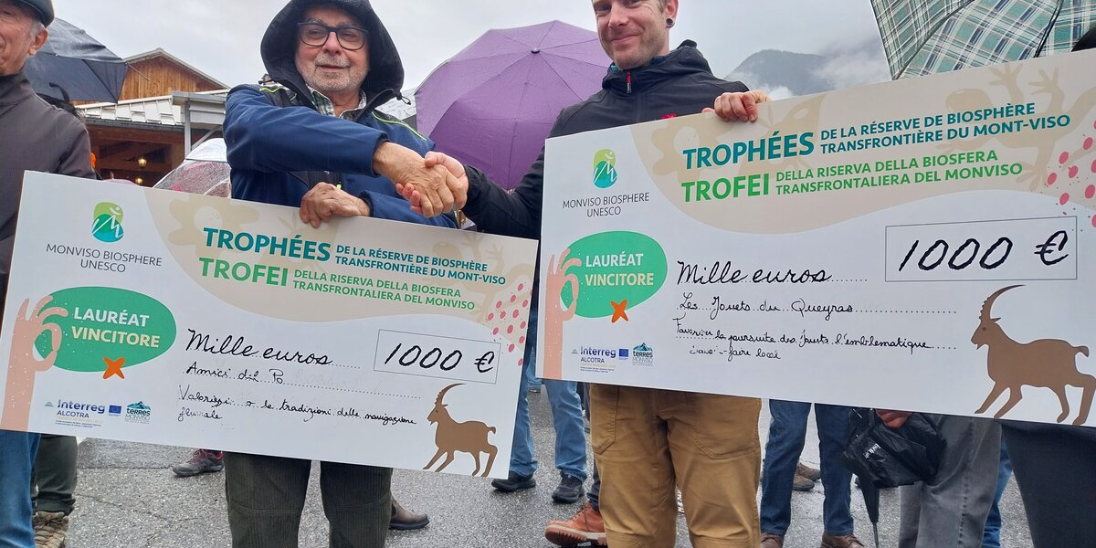 Nell’immagine due uomini sorridenti si stringono la mano all’aperto, sotto la pioggia leggera, entrambi con in mano un grande assegno simbolico da 1.000 euro ciascuno. Gli assegni riportano la scritta “Trophées / Trofei della Riserva della Biosfera transfrontaliera del Monviso – Monviso Biosphere UNESCO”. Sullo sfondo si vedono alcune persone con ombrelli aperti e un paesaggio montano parzialmente coperto dalle nuvole. L’atmosfera è di festa e riconoscimento ufficiale.