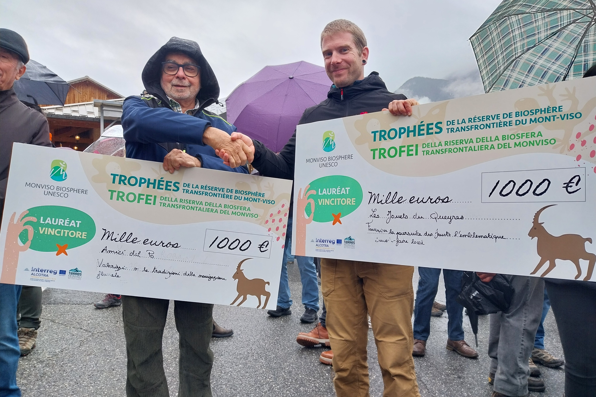 Nell’immagine due uomini sorridenti si stringono la mano all’aperto, sotto la pioggia leggera, entrambi con in mano un grande assegno simbolico da 1.000 euro ciascuno. Gli assegni riportano la scritta “Trophées / Trofei della Riserva della Biosfera transfrontaliera del Monviso – Monviso Biosphere UNESCO”. Sullo sfondo si vedono alcune persone con ombrelli aperti e un paesaggio montano parzialmente coperto dalle nuvole. L’atmosfera è di festa e riconoscimento ufficiale.