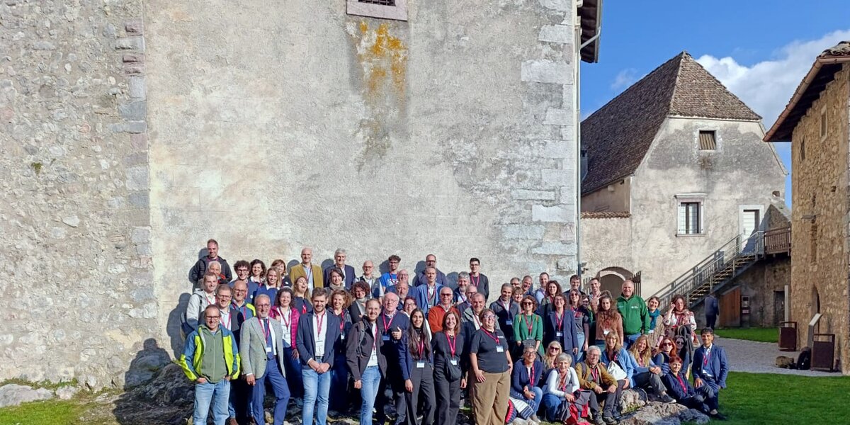 Nell’immagine un grande gruppo di persone posa per una foto di gruppo all’aperto, davanti a un edificio in pietra di aspetto storico. I partecipanti, circa un’ottantina, sono disposti su più file e molti indossano badge o cartellini identificativi al collo, segno di un incontro o convegno ufficiale. Il cielo è sereno e la luce del sole illumina la scena, creando un’atmosfera conviviale e ordinata in un contesto architettonico di pregio.