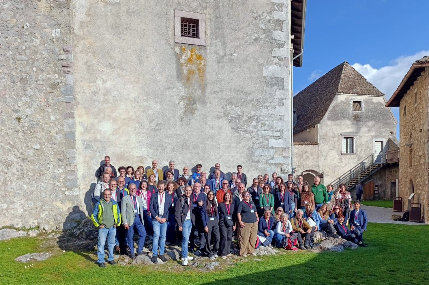 Nell’immagine un grande gruppo di persone posa per una foto di gruppo all’aperto, davanti a un edificio in pietra di aspetto storico. I partecipanti, circa un’ottantina, sono disposti su più file e molti indossano badge o cartellini identificativi al collo, segno di un incontro o convegno ufficiale. Il cielo è sereno e la luce del sole illumina la scena, creando un’atmosfera conviviale e ordinata in un contesto architettonico di pregio.