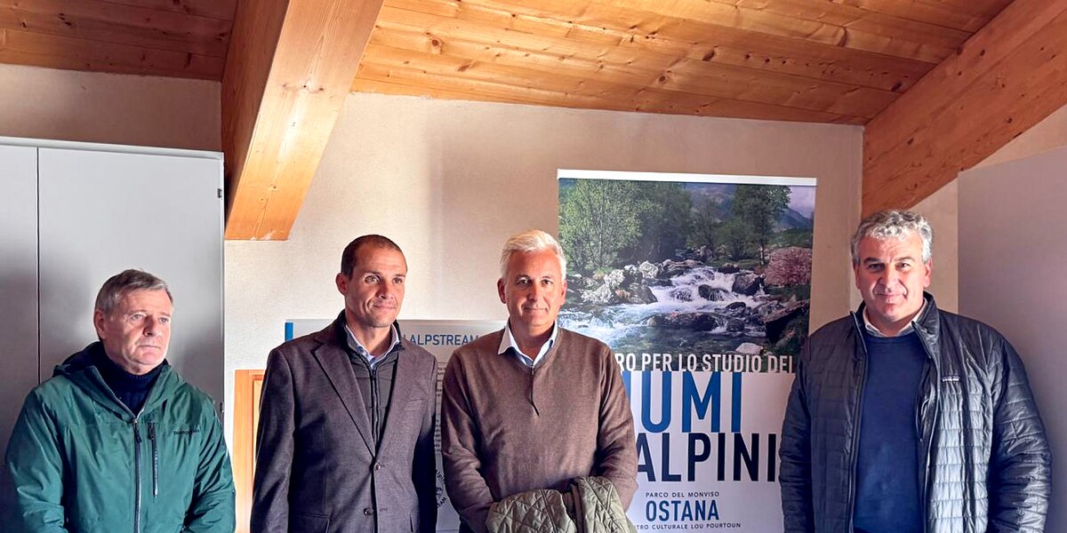 quattro uomini in un ambiente interno con travatura in legno a vista. Si vede un roll up con il logo del Centro per lo Studio dei Fiumi Alpini - ALPSTREAM.