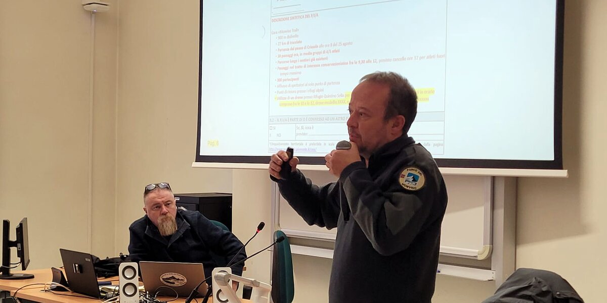 Un tecnico del Parco del Monviso parla al microfono davanti a una classe universitaria, indicando degli appunti proiettati su uno schermo. Accanto a lui, seduto al tavolo con un computer portatile, c’è il professor Stefano Fenoglio che ascolta con attenzione. L’aula è semplice e luminosa, con attrezzatura informatica e materiali di lavoro sul tavolo.