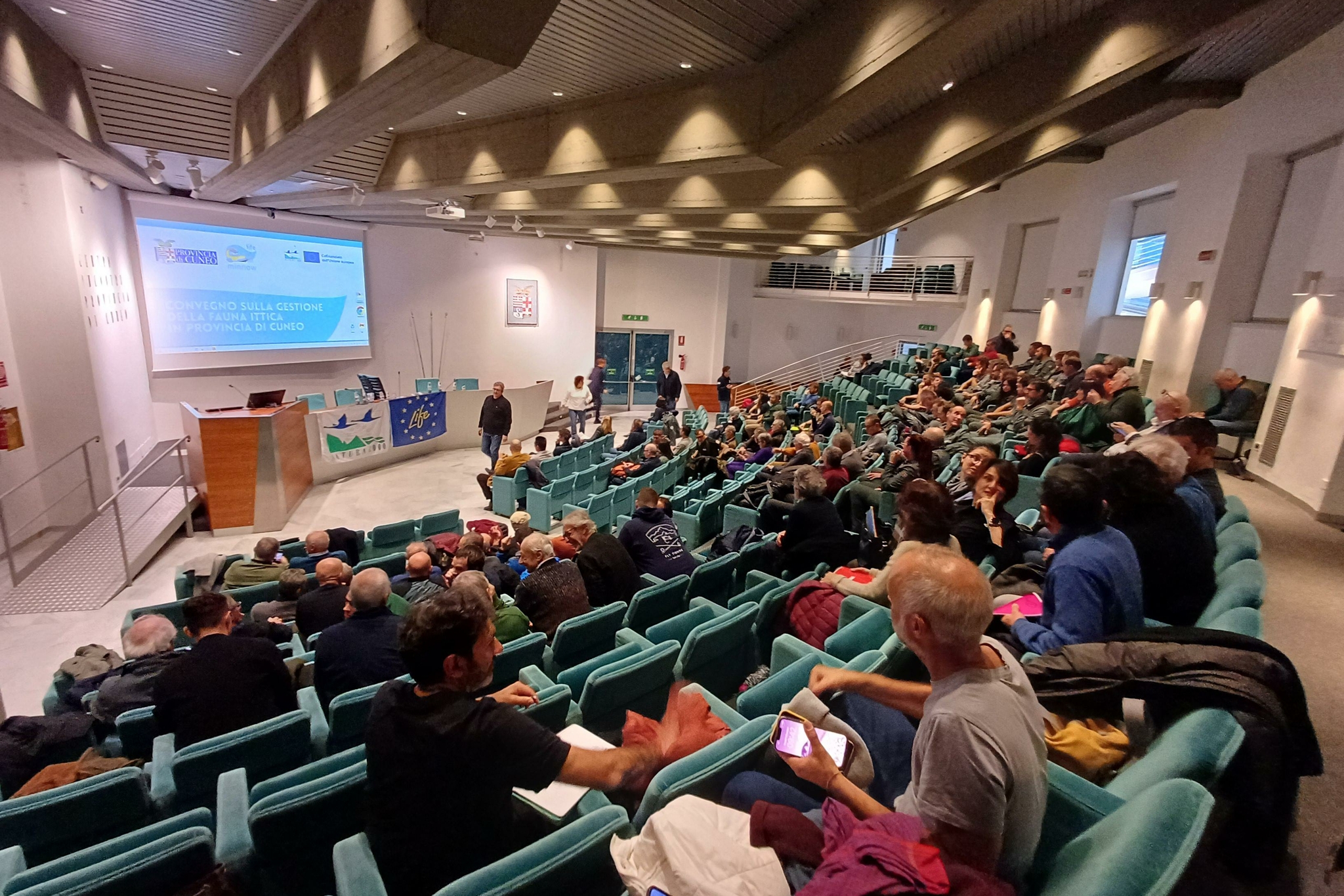 Un’ampia sala conferenze, il Centro Incontri della Provincia di Cuneo, con molte persone sedute su file di poltroncine blu, in attesa dell’inizio di un convegno. Sul palco, davanti a un grande schermo con la scritta “Convegno sulla gestione della fauna ittica in provincia di Cuneo”, alcune persone si muovono tra il tavolo dei relatori e le attrezzature. L’atmosfera è vivace e partecipata, con il pubblico che conversa e prende posto. La sala è luminosa, con soffitto alto e ampie scale laterali.