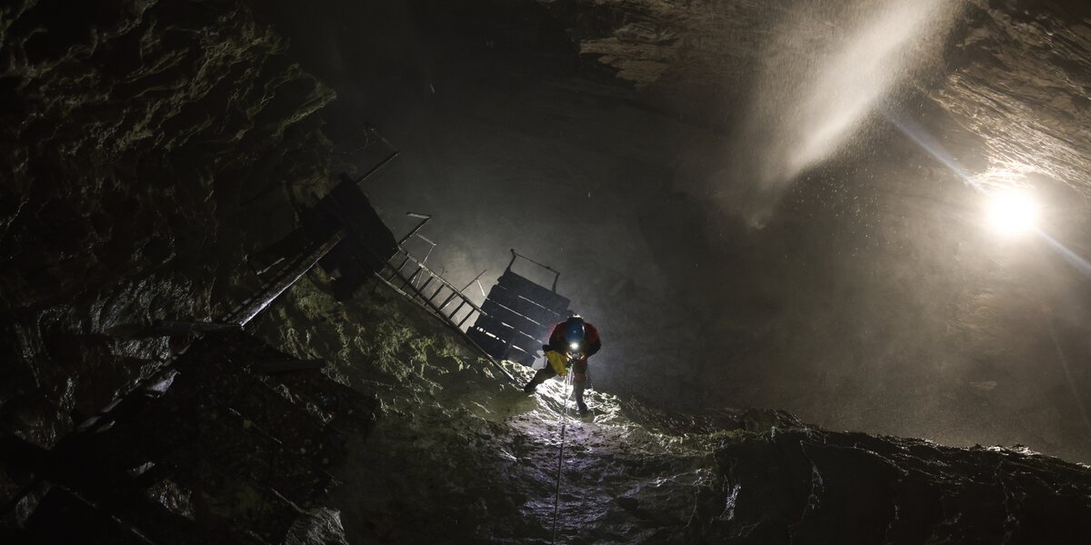 Ambiente sotterraneo scuro e roccioso, la grotta del Rio Martino a Crissolo. Una persona con casco e luce frontale sta risalendo una parete inclinata utilizzando una corda. Alla sua sinistra si intravede una struttura in legno con scale e passerelle, mentre dall’alto a destra entra un fascio di luce che illumina la una cascata di acqua e la roccia, creando un forte contrasto tra buio e chiarore.