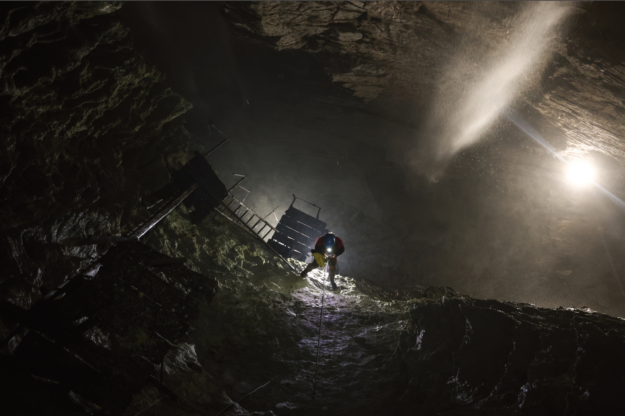 Ambiente sotterraneo scuro e roccioso, la grotta del Rio Martino a Crissolo. Una persona con casco e luce frontale sta risalendo una parete inclinata utilizzando una corda. Alla sua sinistra si intravede una struttura in legno con scale e passerelle, mentre dall’alto a destra entra un fascio di luce che illumina la una cascata di acqua e la roccia, creando un forte contrasto tra buio e chiarore.