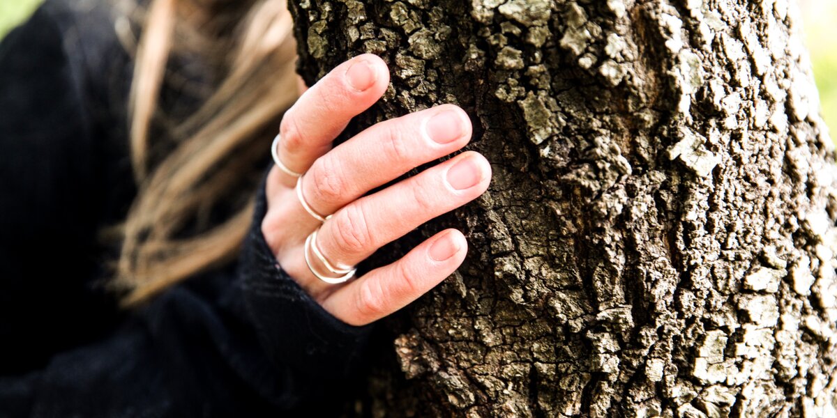 Una mano appoggiata al tronco di un albero, con le dita distese che toccano la corteccia ruvida. La persona indossa un maglione scuro e alcuni anelli sottili alle dita; sullo sfondo si intravedono capelli chiari leggermente sfocati e un ambiente naturale.