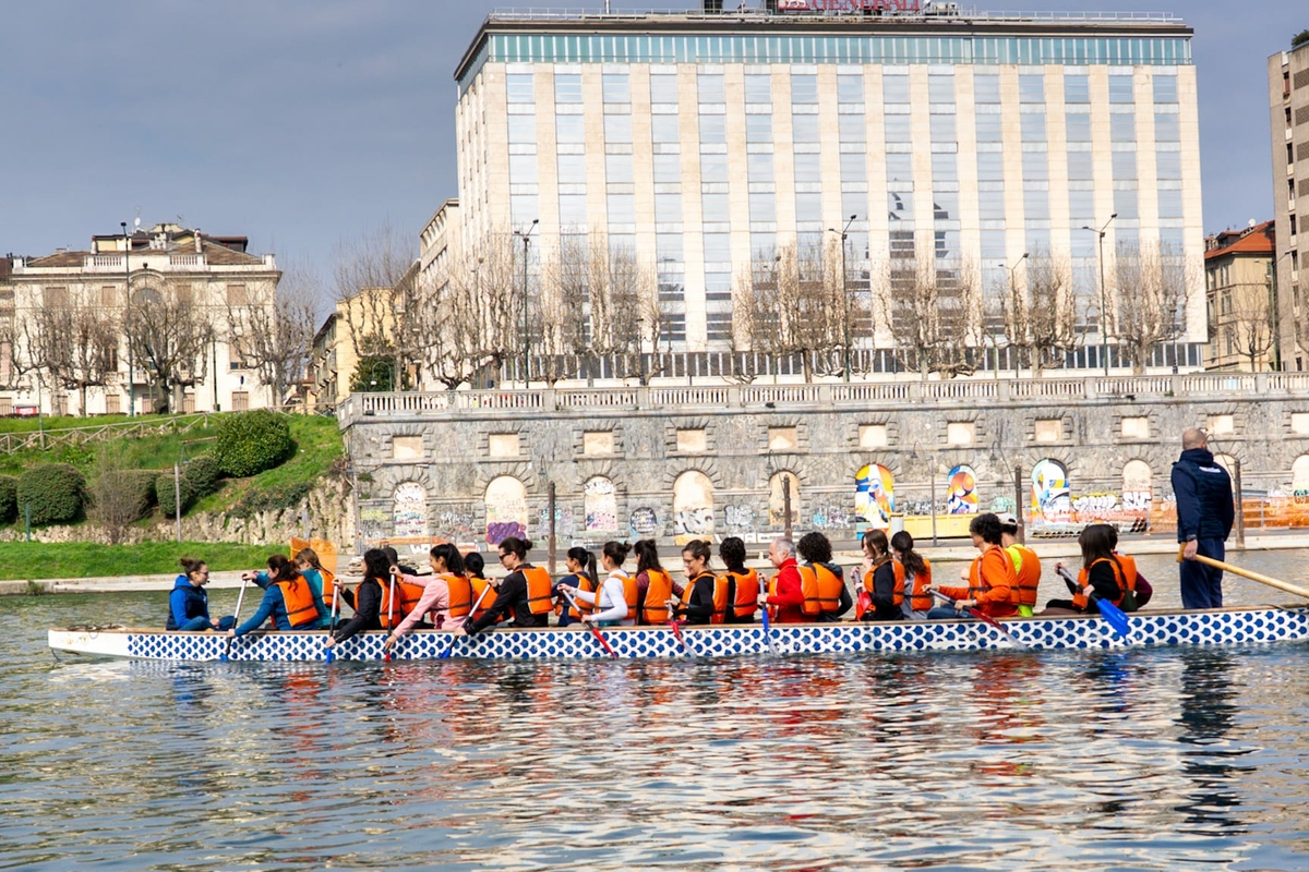 immagine che illustra una giornata di attività sul fiume Po. si osserva una barca piena di cittadini e giovani che pagaiano in un tratto urbano del fiume Po