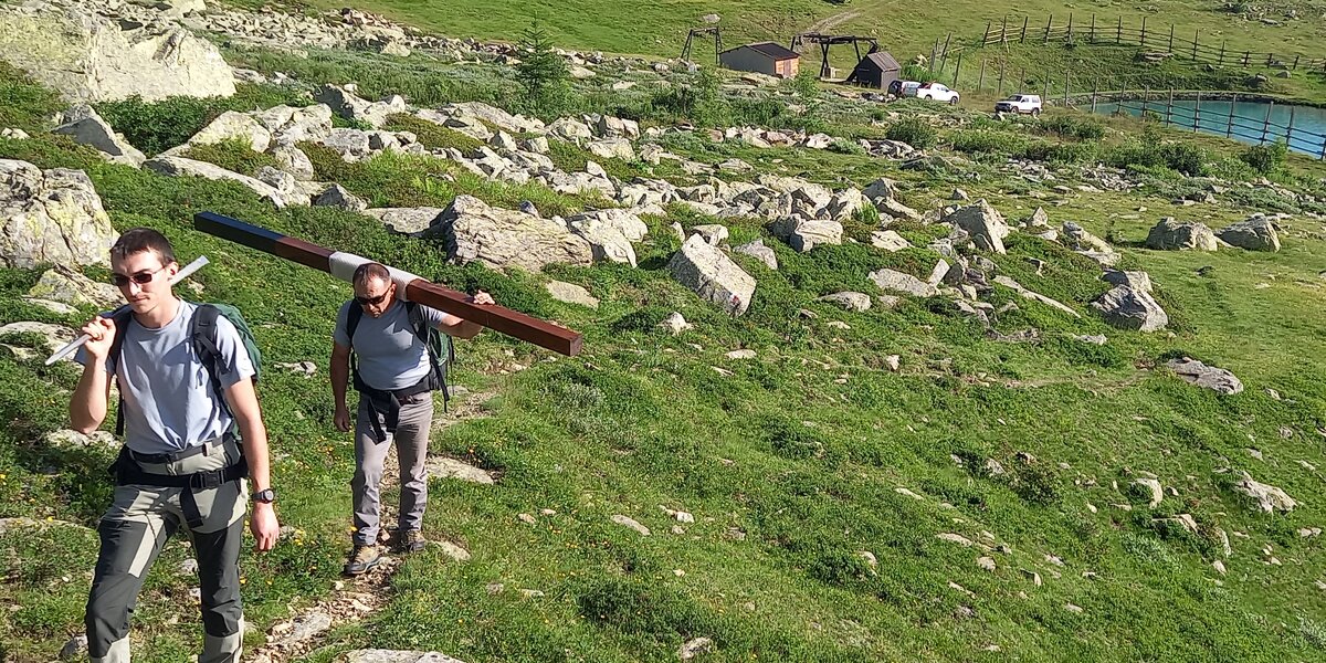 Due operai del Parco del Monviso percorrono un sentiero in ambiente alpino trasportando a spalla una lunga trave di legno e metallo, destinati alla manutenzione della sentieristica. Il sentiero attraversa un pendio erboso disseminato di rocce. Sullo sfondo si vedono alcuni piccoli edifici, alcune automobili parcheggiate e un laghetto artificiale, inseriti in un ampio paesaggio di montagna.