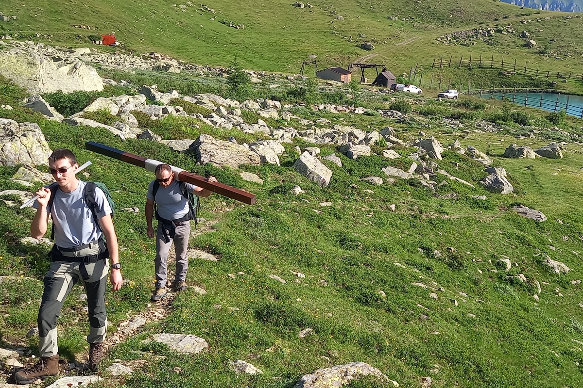 Due operai del Parco del Monviso percorrono un sentiero in ambiente alpino trasportando a spalla una lunga trave di legno e metallo, destinati alla manutenzione della sentieristica. Il sentiero attraversa un pendio erboso disseminato di rocce. Sullo sfondo si vedono alcuni piccoli edifici, alcune automobili parcheggiate e un laghetto artificiale, inseriti in un ampio paesaggio di montagna.