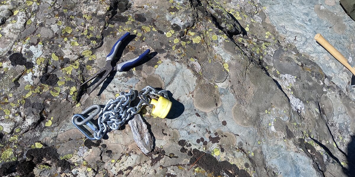 Primo piano di alcuni strumenti da lavoro appoggiati su una grande lastra di roccia in alta montagna: una pinza, un martello e una catena metallica a cui è legata la sonda acquatica in fase di installazione. La superficie della roccia è ricoperta da numerosi licheni di colore giallo e grigio.