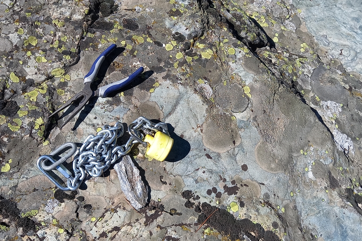 Primo piano di alcuni strumenti da lavoro appoggiati su una grande lastra di roccia in alta montagna: una pinza, un martello e una catena metallica a cui è legata la sonda acquatica in fase di installazione. La superficie della roccia è ricoperta da numerosi licheni di colore giallo e grigio.