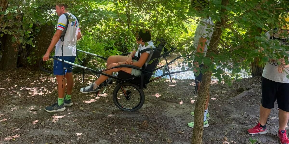Un gruppo di quattro persone su un sentiero boscoso, dove una donna siede su una speciale sedia a rotelle fuoristrada monoruota, pronta per essere trasportata dagli altri membri del gruppo.