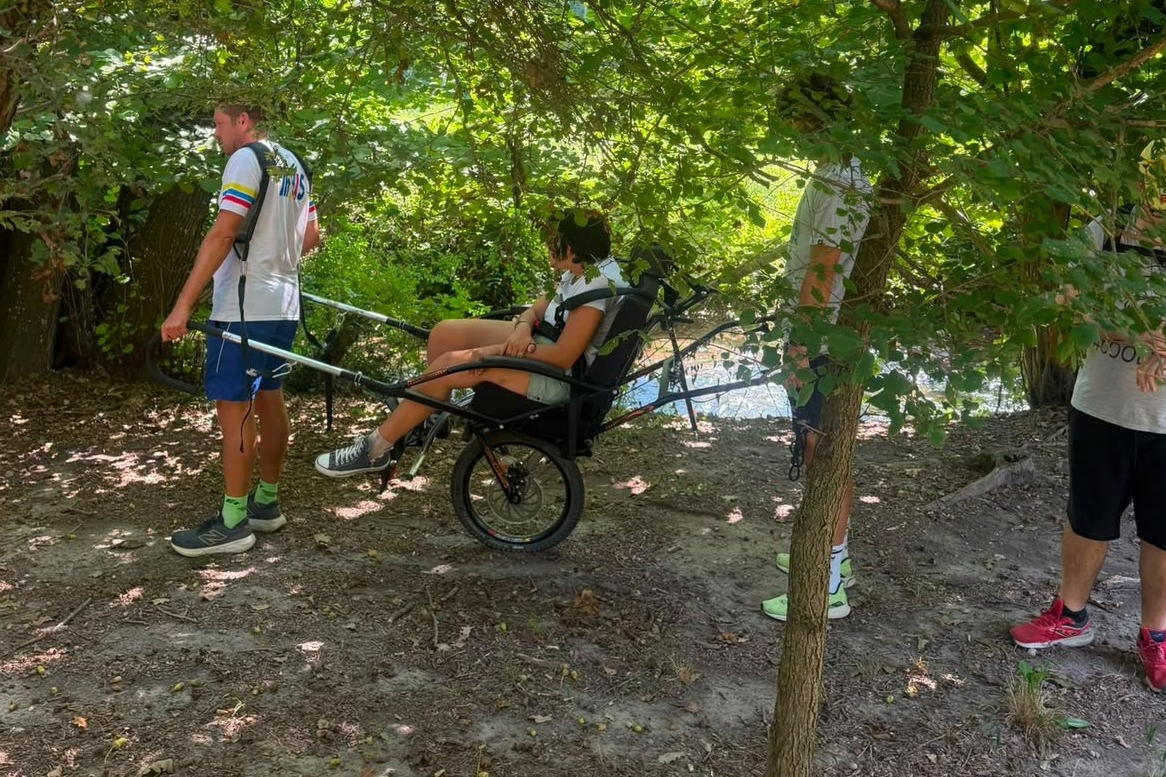 Un gruppo di quattro persone su un sentiero boscoso, dove una donna siede su una speciale sedia a rotelle fuoristrada monoruota, pronta per essere trasportata dagli altri membri del gruppo.