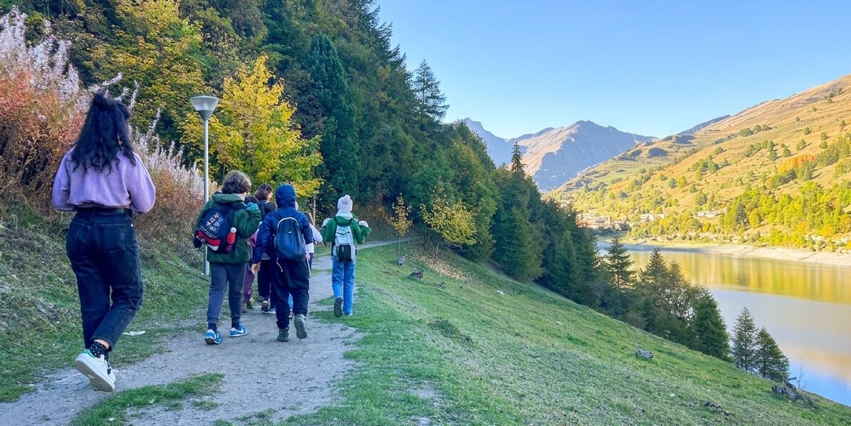 Una fotografia panoramica a colori scattata da dietro un gruppo di sei escursionisti che percorrono un sentiero di ghiaia su un pendio erboso. La donna in primo piano sulla sinistra indossa un maglione viola e jeans scuri, con una borsa a tracolla di tela. Davanti a lei ci sono cinque persone con zaini, che camminano in fila sul sentiero. Sulla sinistra, una fitta foresta mista di alberi decidui e conifere con fogliame autunnale verde e giallo si estende su per il pendio. Sulla destra, un pendio erboso scende verso un lago calmo che riflette il cielo blu limpido. Le montagne si ergono maestose sullo sfondo, con pendii rocciosi e coperti di vegetazione. La luce del sole pomeridiano illumina la scena.