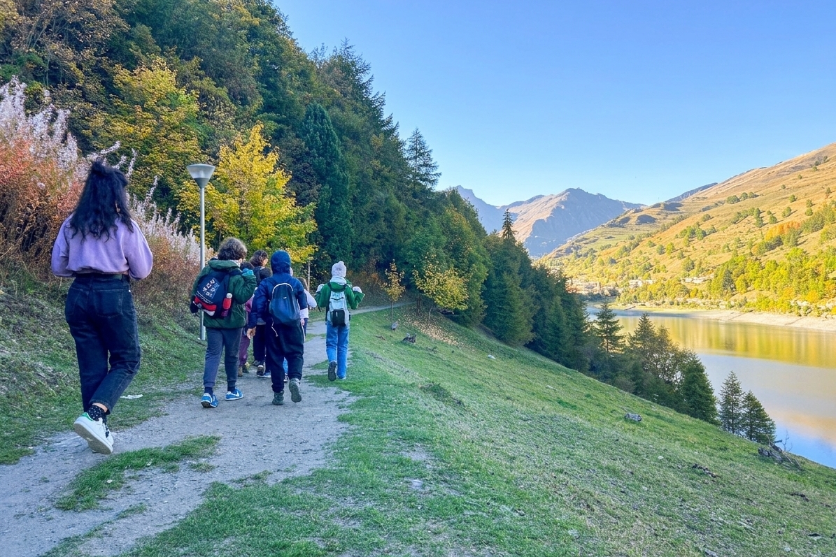 Una fotografia panoramica a colori scattata da dietro un gruppo di sei escursionisti che percorrono un sentiero di ghiaia su un pendio erboso. La donna in primo piano sulla sinistra indossa un maglione viola e jeans scuri, con una borsa a tracolla di tela. Davanti a lei ci sono cinque persone con zaini, che camminano in fila sul sentiero. Sulla sinistra, una fitta foresta mista di alberi decidui e conifere con fogliame autunnale verde e giallo si estende su per il pendio. Sulla destra, un pendio erboso scende verso un lago calmo che riflette il cielo blu limpido. Le montagne si ergono maestose sullo sfondo, con pendii rocciosi e coperti di vegetazione. La luce del sole pomeridiano illumina la scena.