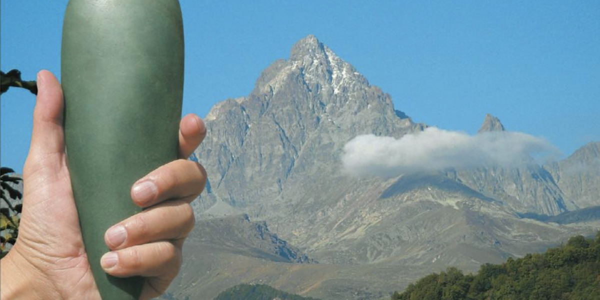 Una mano tiene un grande e liscio oggetto verde a forma di goccia, un'ascia in giada. Sullo sfondo, un maestoso paesaggio montano sotto un cielo blu con la cima di una montagna rocciosa, il Monviso.