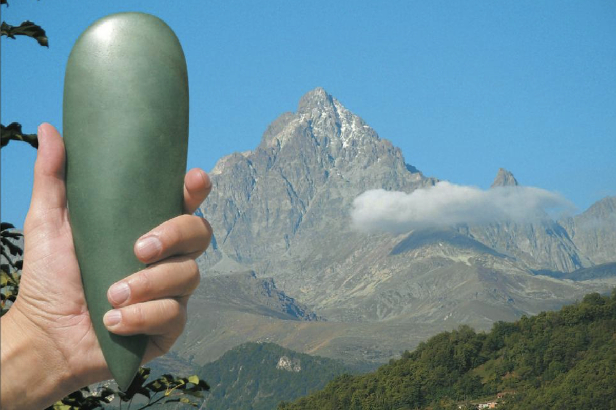 Una mano tiene un grande e liscio oggetto verde a forma di goccia, un'ascia in giada. Sullo sfondo, un maestoso paesaggio montano sotto un cielo blu con la cima di una montagna rocciosa, il Monviso.