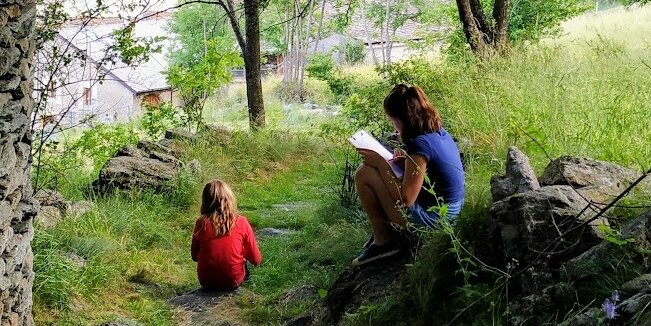 Due bambine sedute all'aperto tra alberi e muretti a secco: una di spalle con maglia rossa e una di profilo che scrive su un blocco per appunti.