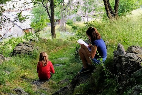 Due bambine sedute all'aperto tra alberi e muretti a secco: una di spalle con maglia rossa e una di profilo che scrive su un blocco per appunti.