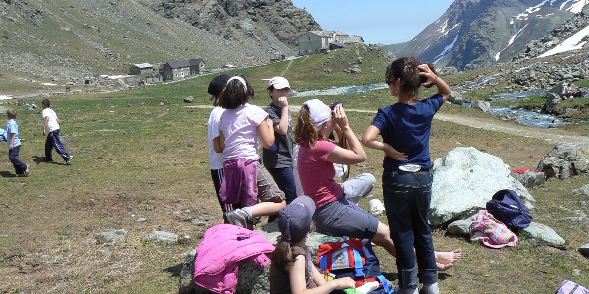 Un gruppo di bambini fa una sosta durante un'escursione a pian del re, con zaini colorati tra le rocce e costruzioni in pietra sullo sfondo.