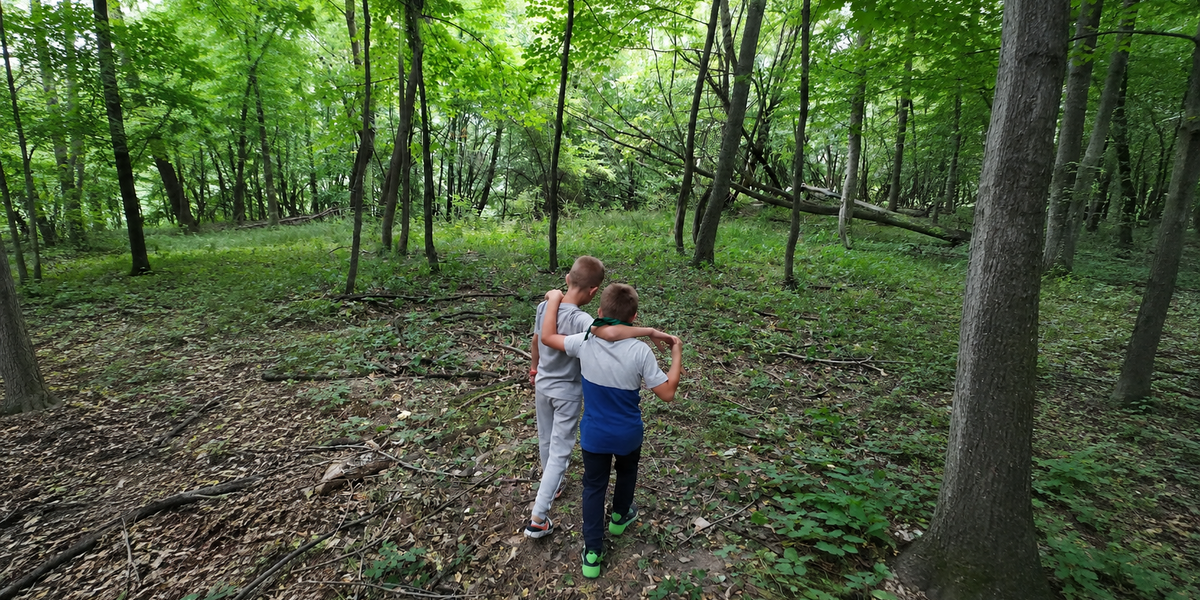 Due bambini visti di spalle camminano abbracciati su un sentiero sterrato all'interno di un bosco rigoglioso.