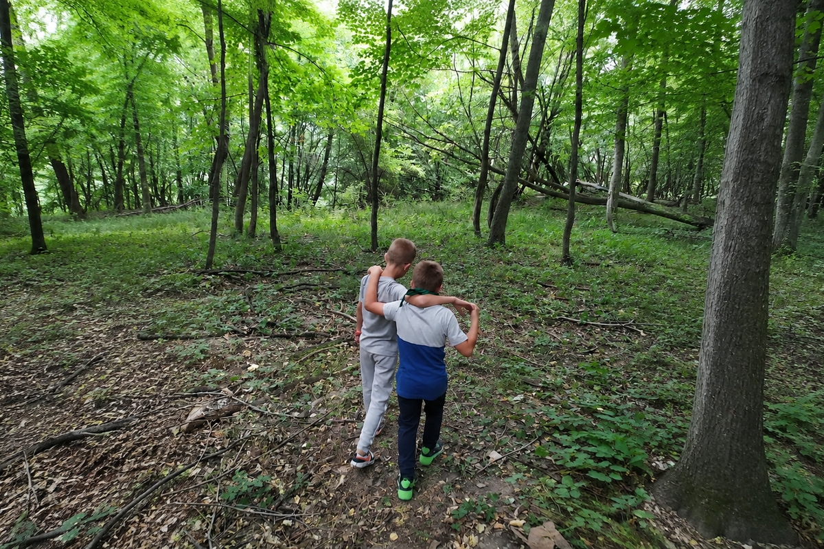 Due bambini visti di spalle camminano abbracciati su un sentiero sterrato all'interno di un bosco rigoglioso.