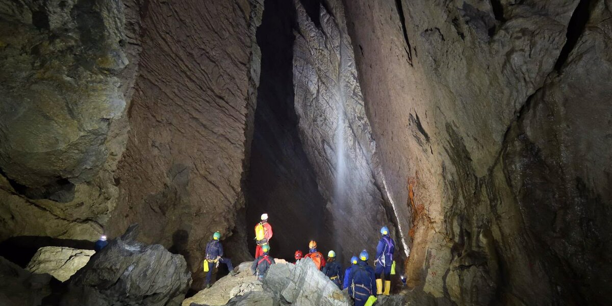Gruppo di visitatori con caschi e luci frontali all'interno di un'alta grotta di pietra, mentre osservano una cascata sotterranea.