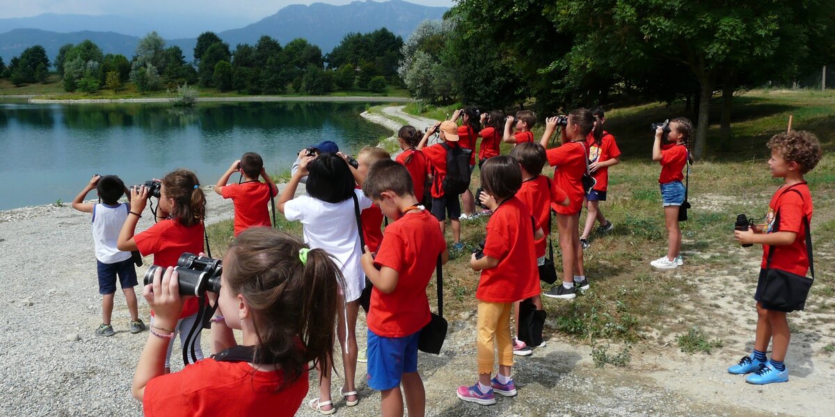 attività di educazione ambientale con bambine e bambini all'aperto che guardano con un binocolo verso un lago. tutti indossano una maglietta rossa.