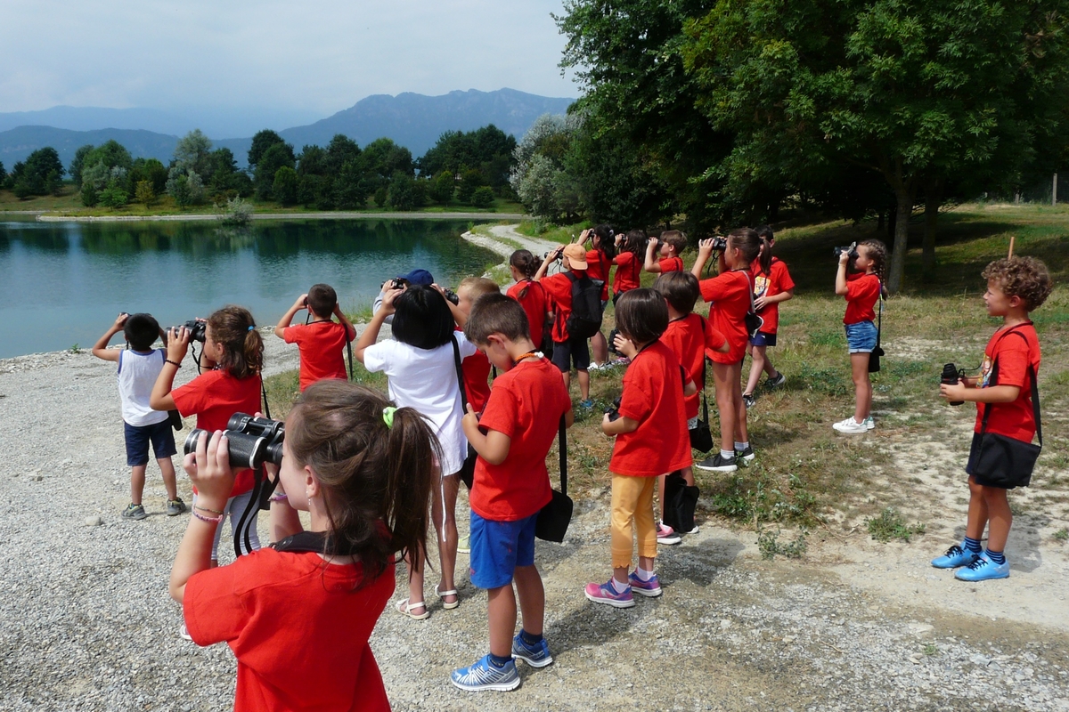 attività di educazione ambientale con bambine e bambini all'aperto che guardano con un binocolo verso un lago. tutti indossano una maglietta rossa.