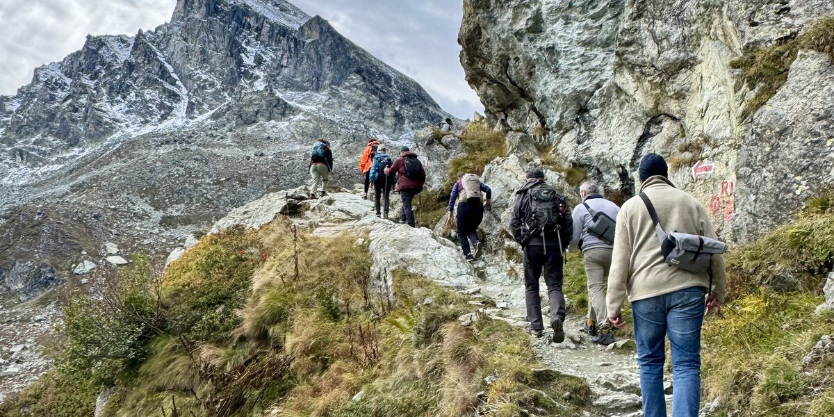 Un gruppo di sette escursionisti con zaini sale su un sentiero di montagna roccioso e ripido, con un'imponente cima innevata sullo sfondo sotto un cielo nuvoloso. Il percorso è fiancheggiato da pareti di roccia e vegetazione autunnale.