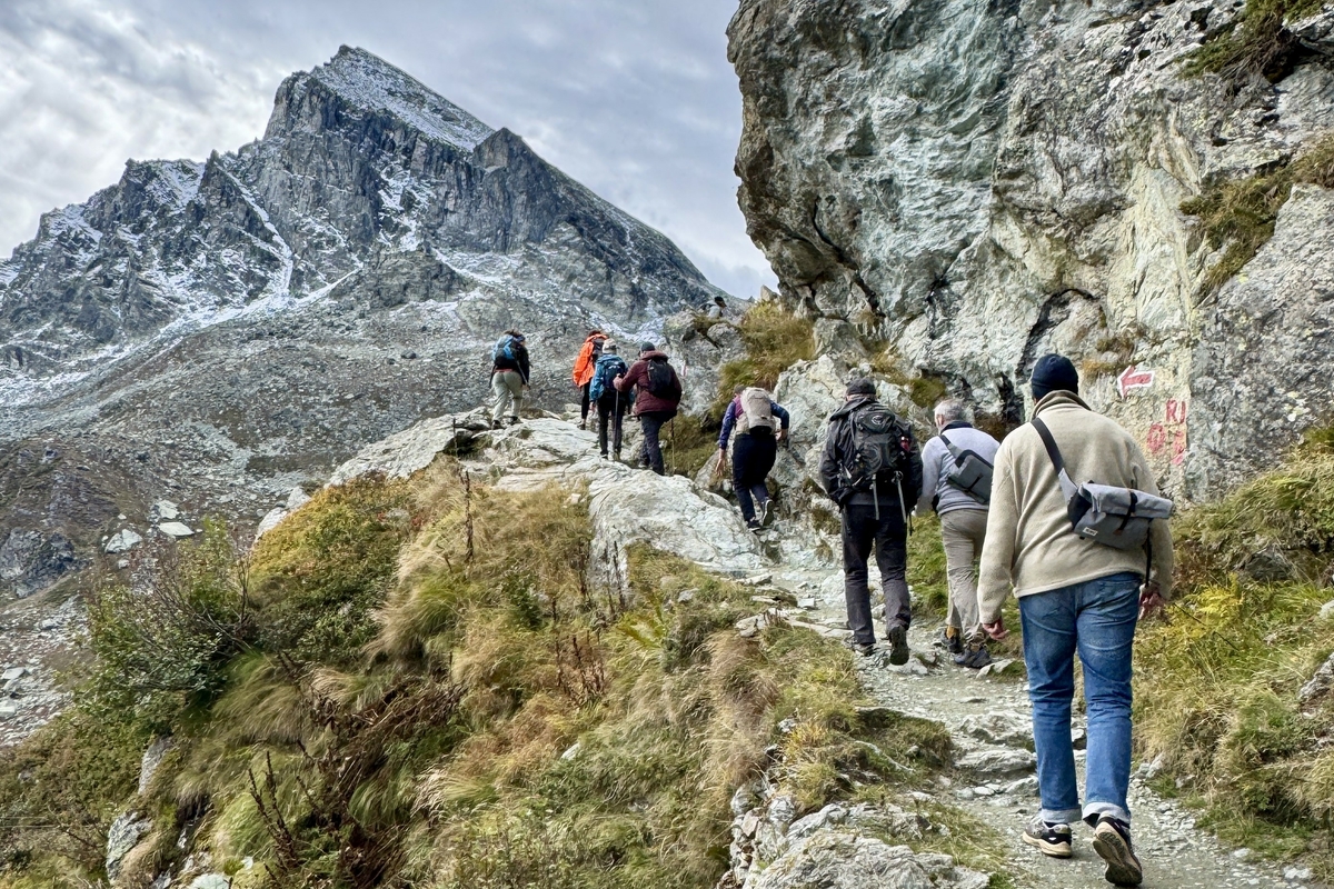 Un gruppo di sette escursionisti con zaini sale su un sentiero di montagna roccioso e ripido, con un'imponente cima innevata sullo sfondo sotto un cielo nuvoloso. Il percorso è fiancheggiato da pareti di roccia e vegetazione autunnale.
