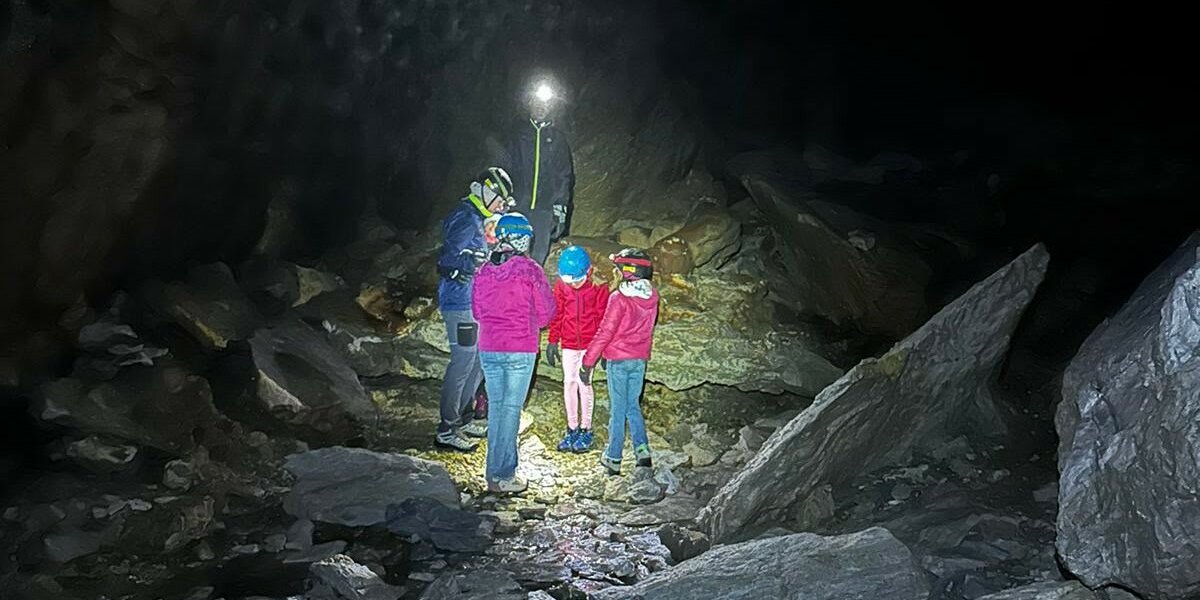Un gruppo di persone, tra cui alcuni bambini con caschi protettivi e torce frontali illuminate, si trova all'interno di una grotta buia circondata da grandi roccia ed elementi rocciosi.