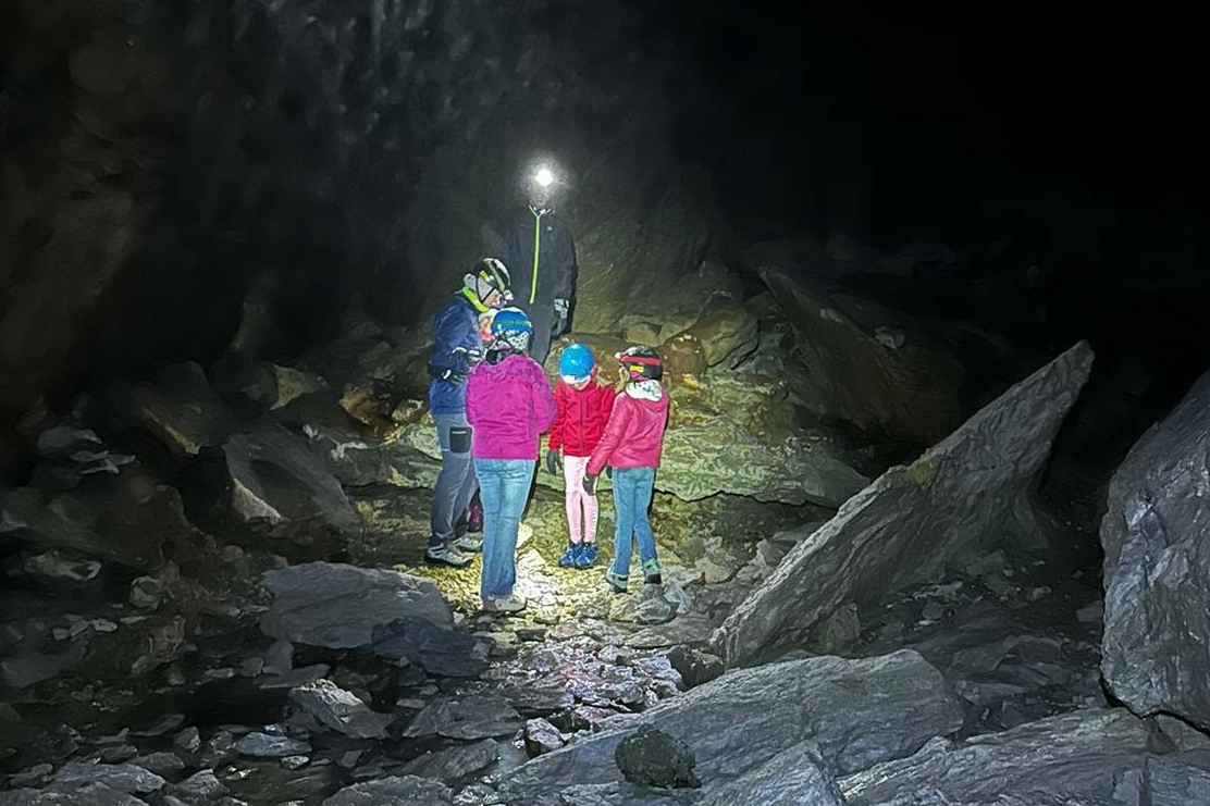 Un gruppo di persone, tra cui alcuni bambini con caschi protettivi e torce frontali illuminate, si trova all'interno di una grotta buia circondata da grandi roccia ed elementi rocciosi.