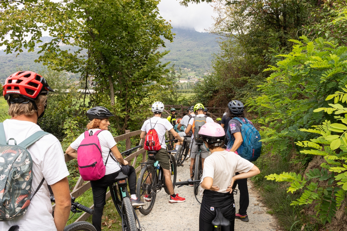 Un gruppo di ciclisti con caschi e zaini è fermo lungo un sentiero sterrato immerso nel verde, affacciato su un paesaggio collinare e montuoso sullo sfondo.