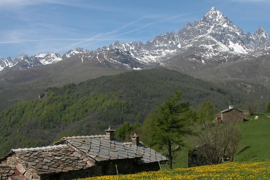 Primo piano di una tradizionale baita di montagna costruita in pietra e con un tetto spiovente in tegole di ardesia, situata in un alpeggio verde. Dietro la baita si estende una vasta valle boscosa e l'imponente catena delle Alpi, con cime innevate e rocciose e il Monviso sulla destra.