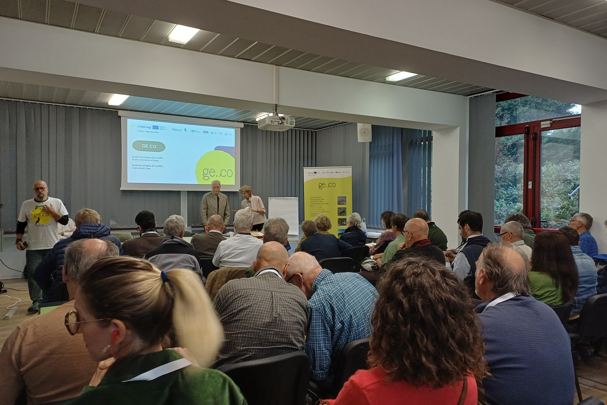 **Testo alternativo (alt text):**  Numerose persone partecipano a un incontro pubblico in una sala conferenze, sedute di fronte ai relatori. Sul fondo, uno schermo proietta una presentazione del progetto GE.CO, affiancato da un pannello informativo con la stessa grafica. Alcuni relatori sono in piedi nella parte anteriore della sala.
